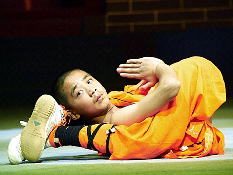 A young martial artist in an orange uniform performs a flexibility pose with one leg behind his head.
